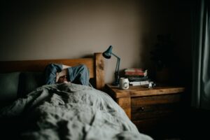 A person rests under blankets in a dimly lit, peaceful bedroom with closed curtains and a small lamp on a nightstand.