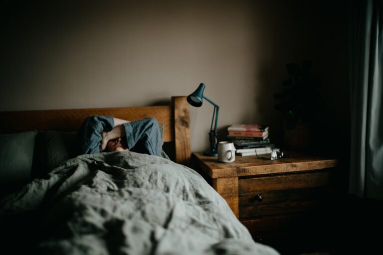 A person rests under blankets in a dimly lit, peaceful bedroom with closed curtains and a small lamp on a nightstand.